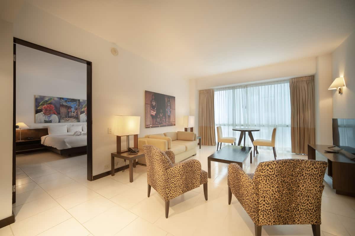 A hotel room with a bed, a sofa, two leopard print armchairs, a coffee table, a TV, and a dining table with chairs.