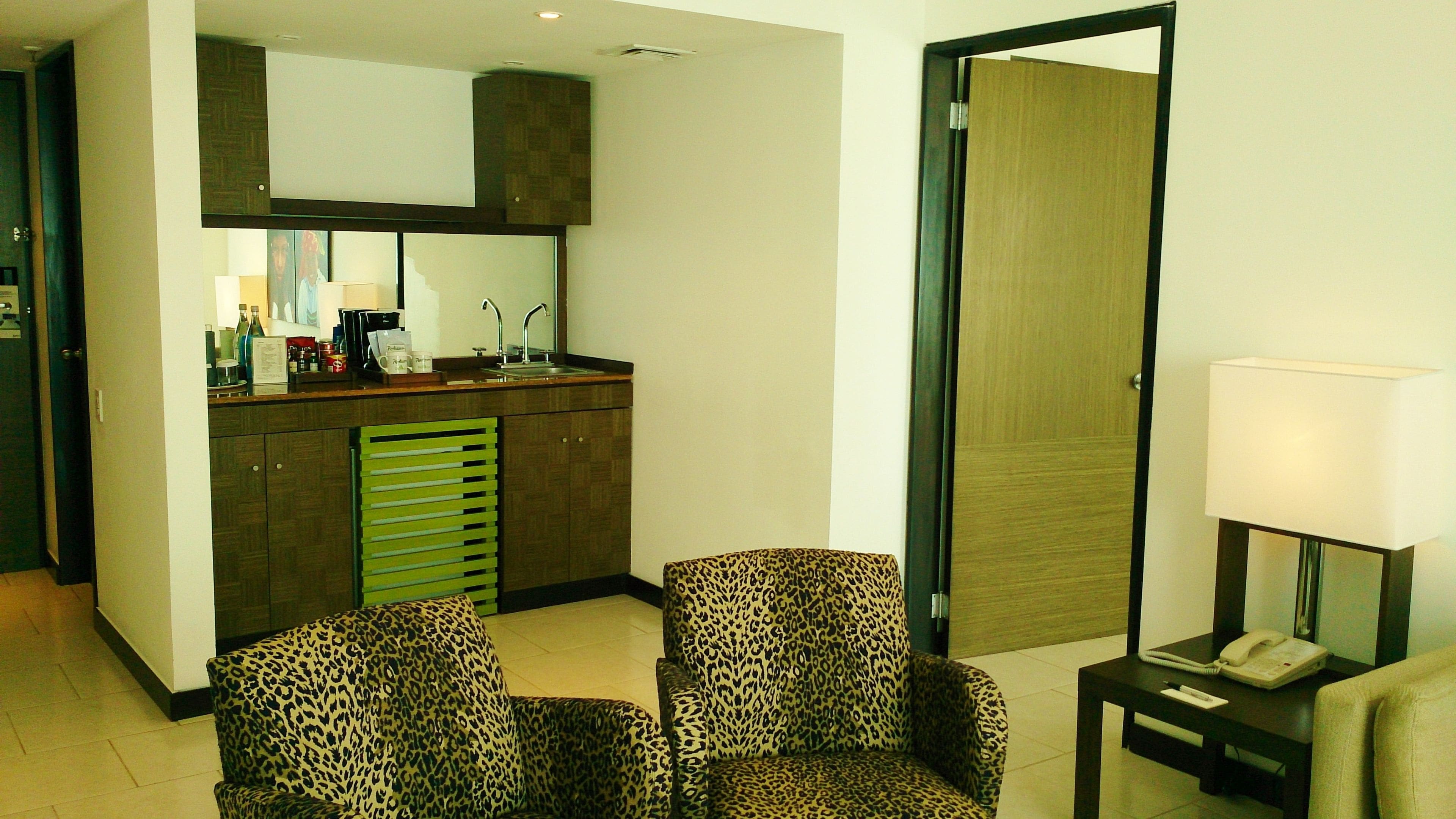 A hotel room with a kitchenette, two leopard-print armchairs, a bedside table with a lamp, and a door leading to another room.