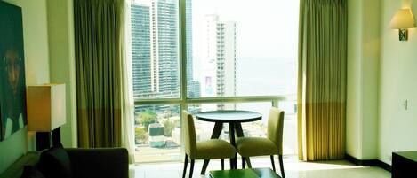 A hotel room with a large window, two chairs, a small table, and a lamp.