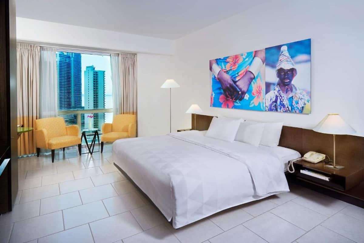 A hotel room with a large bed, two chairs, a desk, and a large window with a city view.