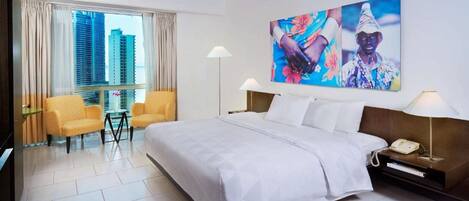 Habitación de hotel con una cama grande, dos sillones, un escritorio y un ventanal con vistas a la ciudad.