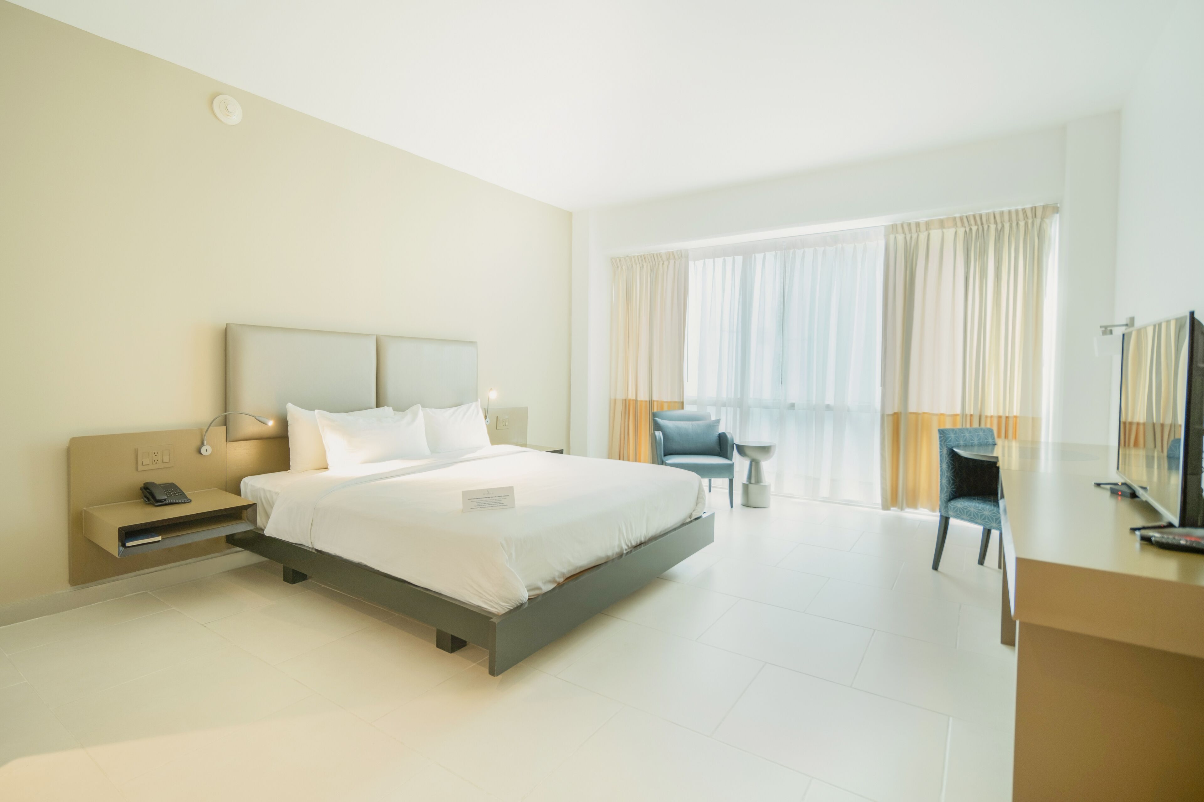 A modern hotel room with a large bed, bedside table, lamp, chair, and a desk with a TV.
