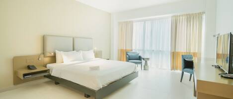 A modern hotel room with a large bed, bedside table, lamp, chair, and a desk with a TV.