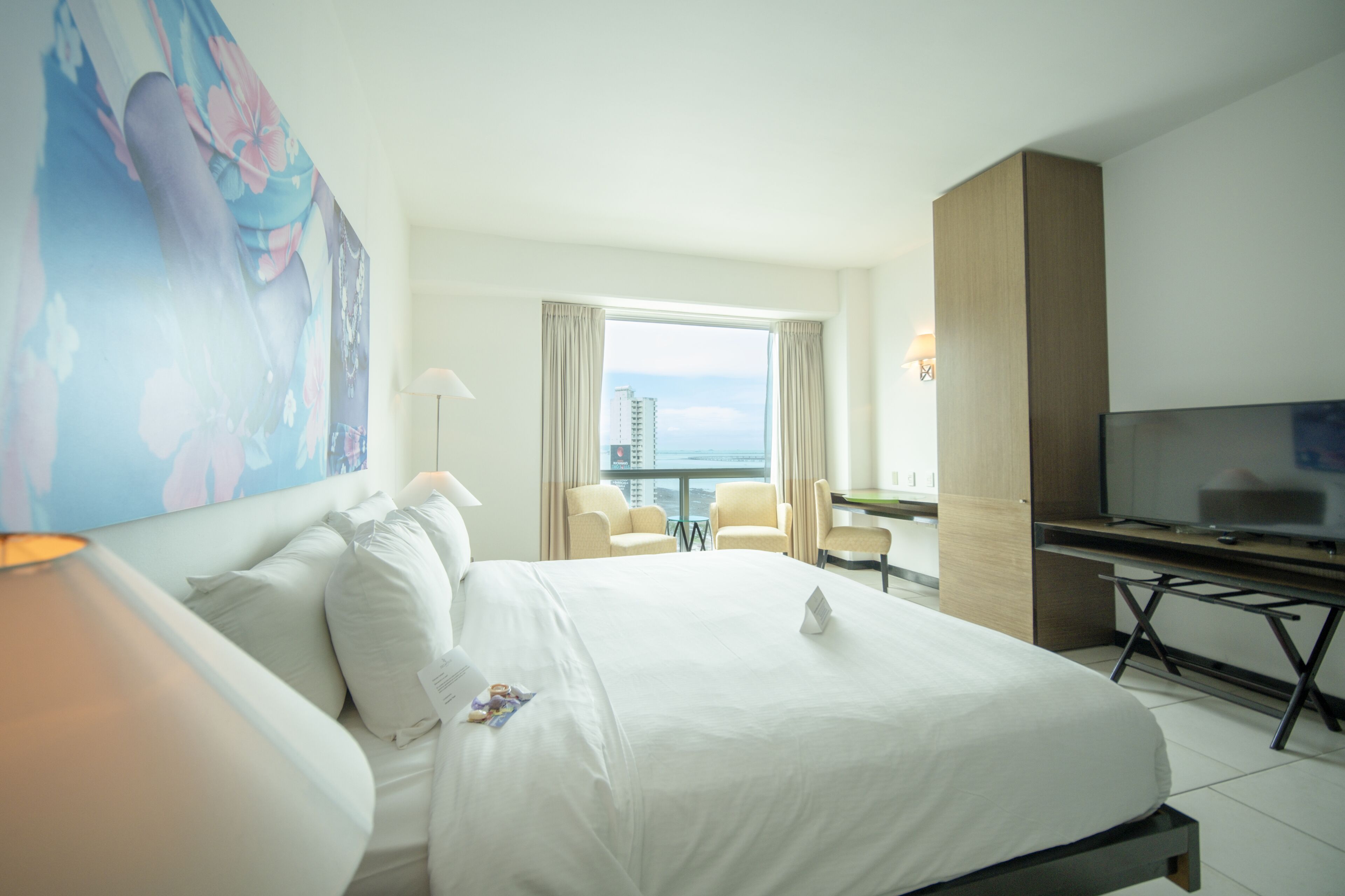 A hotel room with a large bed, a desk, a TV, and a view of the city.