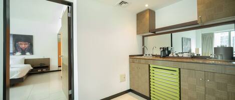 A modern hotel room with a kitchenette, a bed with a headboard, and a large mirror above the counter.