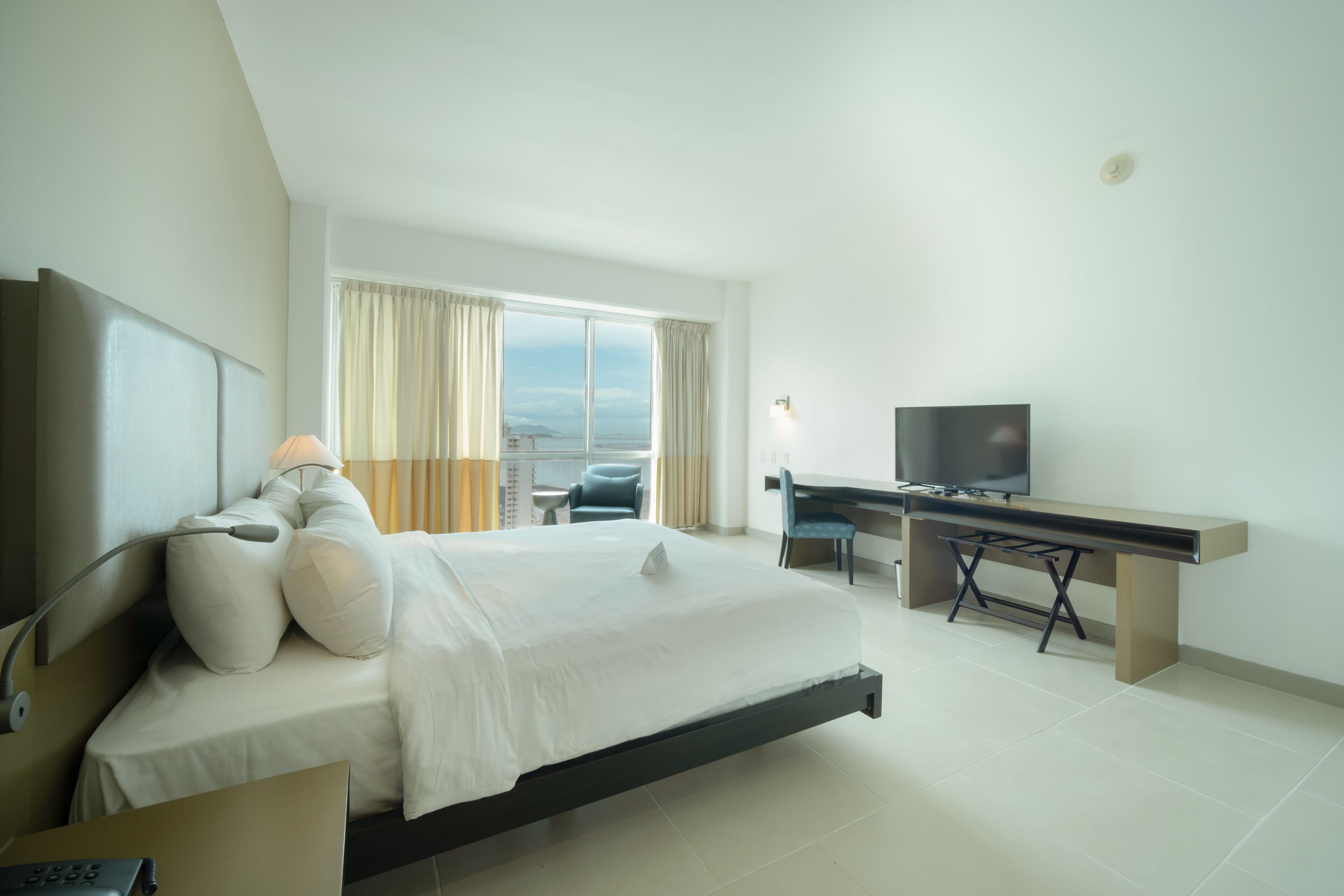 A modern hotel room with a bed, desk, TV, and a view of the city.