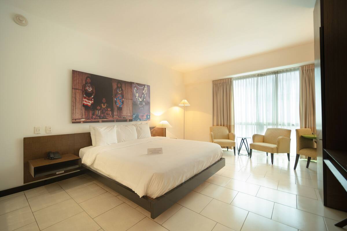 A hotel room with a large bed, two bedside tables, a desk, two chairs, a painting on the wall, and a window with curtains.