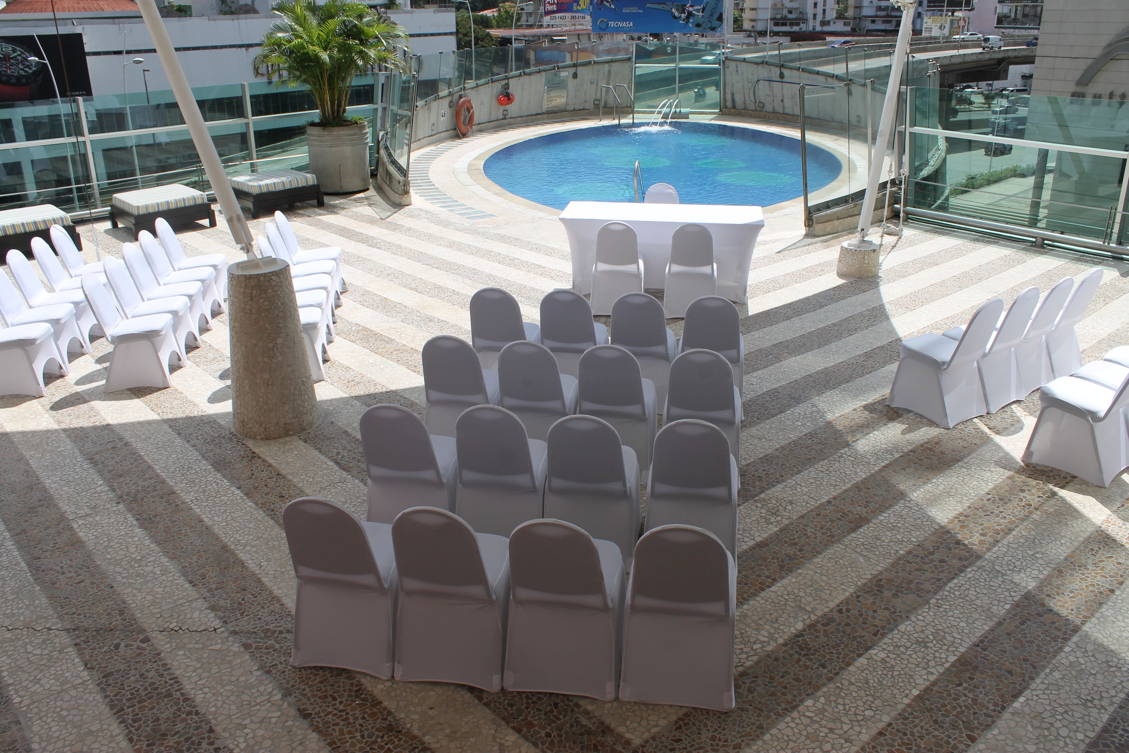 A rooftop pool area with white lounge chairs arranged in rows, a central table, and a glass railing overlooking the pool.
