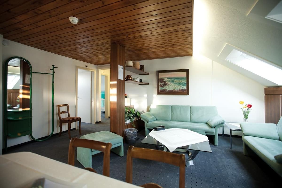Ein gemütliches Zimmer mit Holzboden, einem grünen Sofa, einem Holzstuhl, einem kleinen Tisch mit weißer Tischdecke und einem Bild an der Wand.
