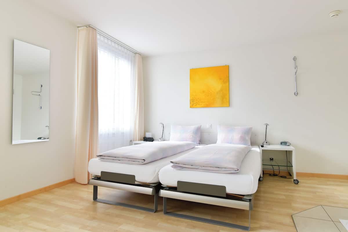 Ein modernes Hotelzimmer mit zwei Betten, einem großen Spiegel, einem Nachttisch mit Lampe und einem Fenster mit Vorhängen.