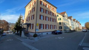 Un hôtel à plusieurs étages, doté d’une façade beige et de volets rouges, situé à l’angle d’une rue, avec un ciel bleu dégagé.