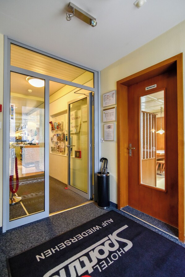 A hotel entrance with a reception desk, a glass door leading inside, and a wooden door labeled 'Restaurant'.
