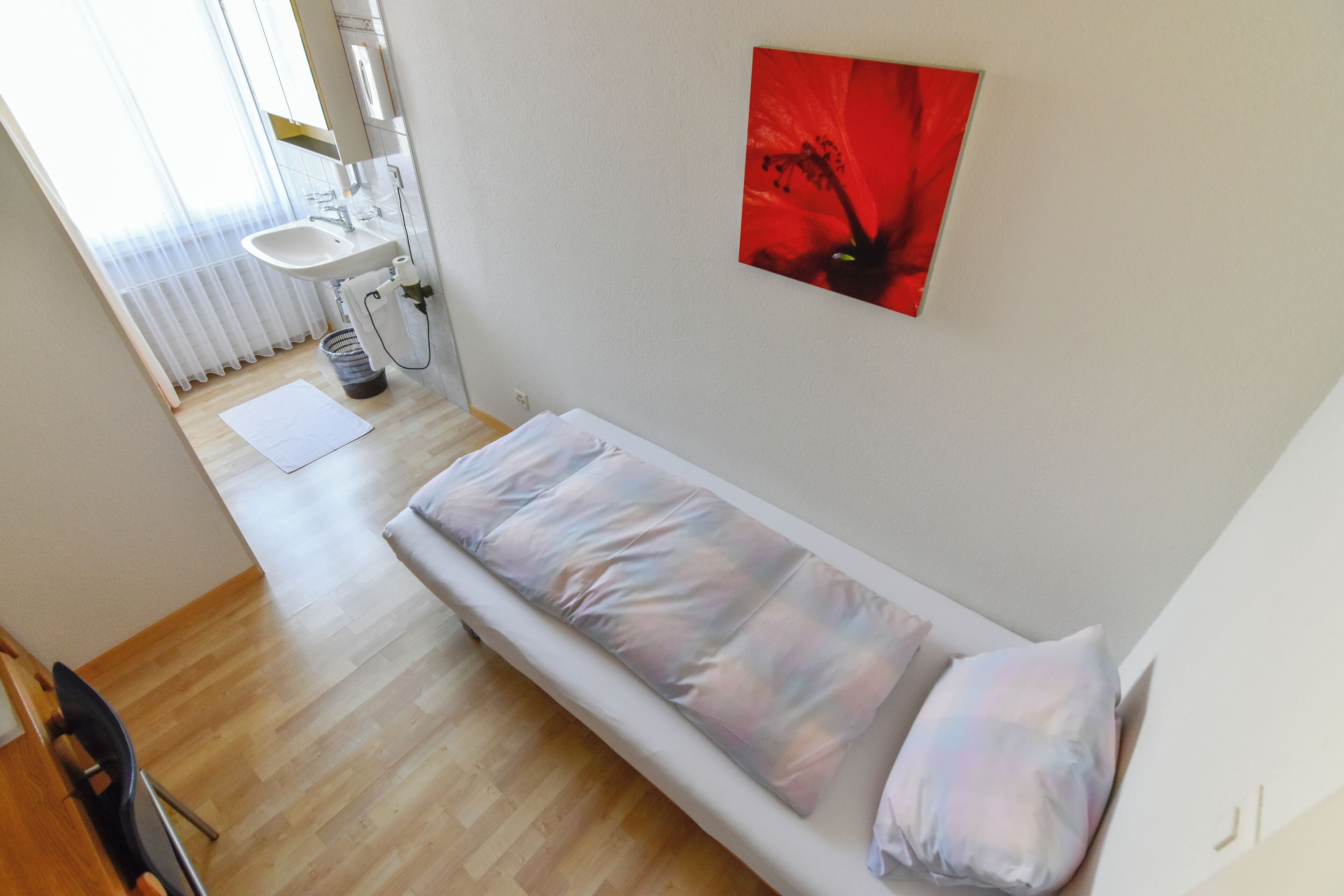 A small, single-bed room with a white bed, a red flower painting on the wall, a white sink, and a window with sheer curtains.