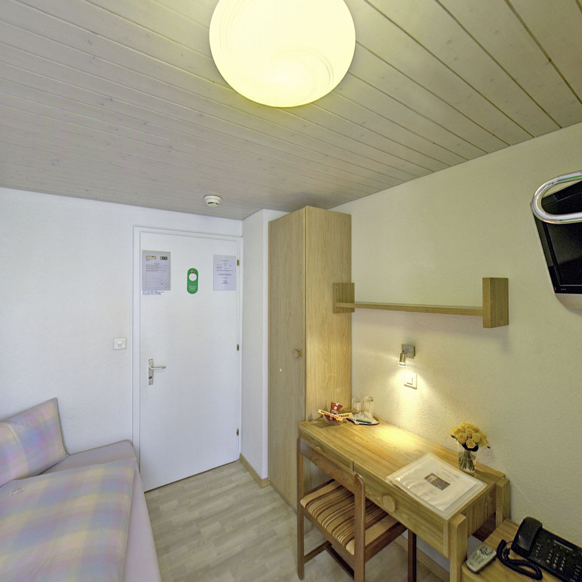 Una habitación pequeña y ordenada, con cama, escritorio con silla, televisor de pared y luz en el techo.
