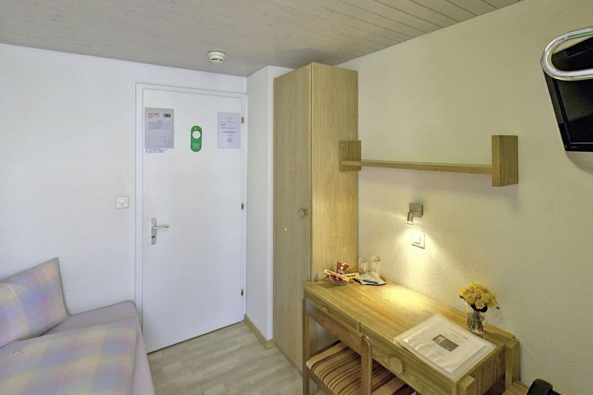 Ein kleiner, ordentlicher Raum mit einem Bett, einem Schreibtisch mit Stuhl, einem Wand-Fernseher und einer Deckenleuchte.
