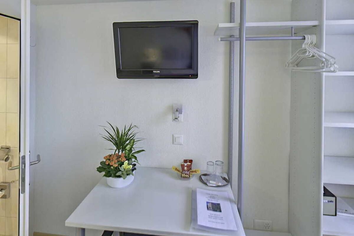 Ein kleines Zimmer mit einem wandmontierten Flachbildfernseher, einem weißen Tisch mit einer Topfpflanze, einer Ablage mit einem Glas und einer Packung sowie einer weißen Schrankwand mit Kleiderhaken.