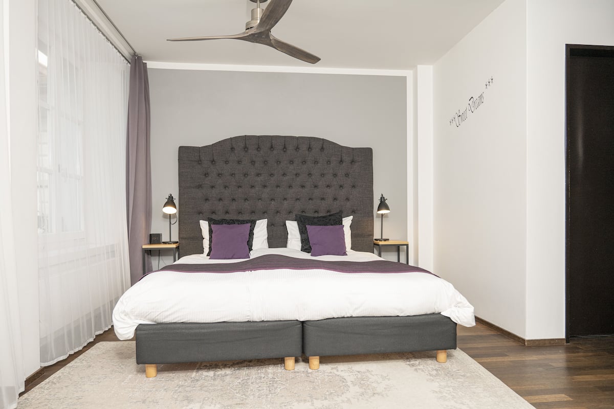 A modern bedroom with a tufted headboard, a large bed with a white and purple bedding set, two bedside tables with lamps, and a ceiling fan.