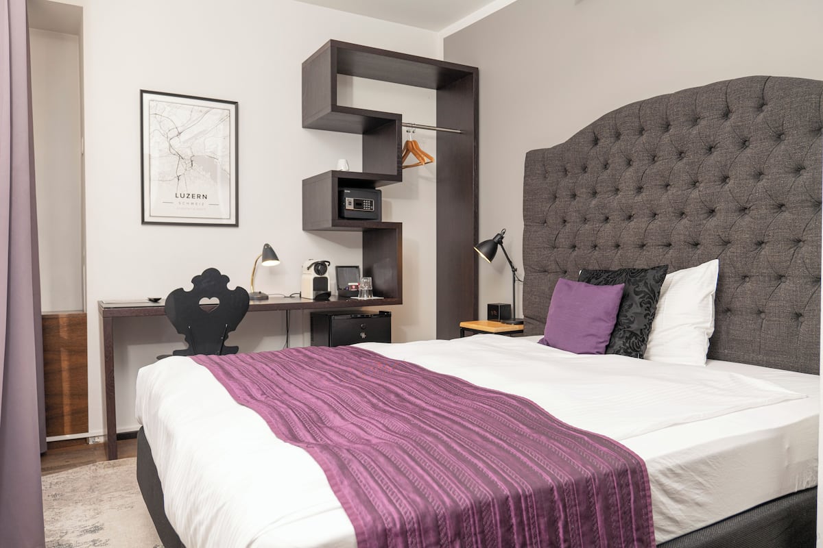 A modern hotel room with a large bed, a desk, and a chair. There is a map on the wall and a small shelf with a radio and a clock.