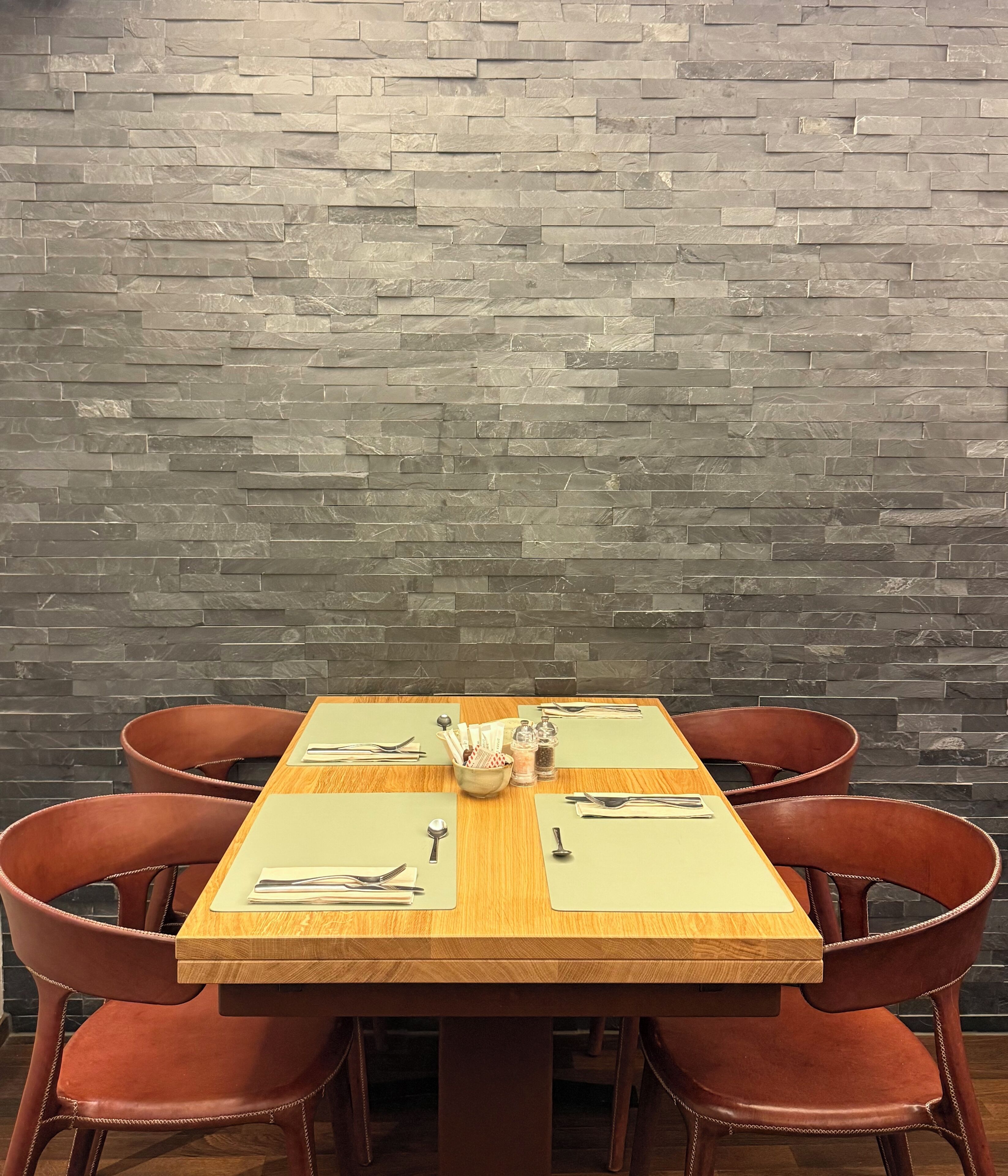 A dining table with four chairs, set with cutlery and condiments, against a stone wall.