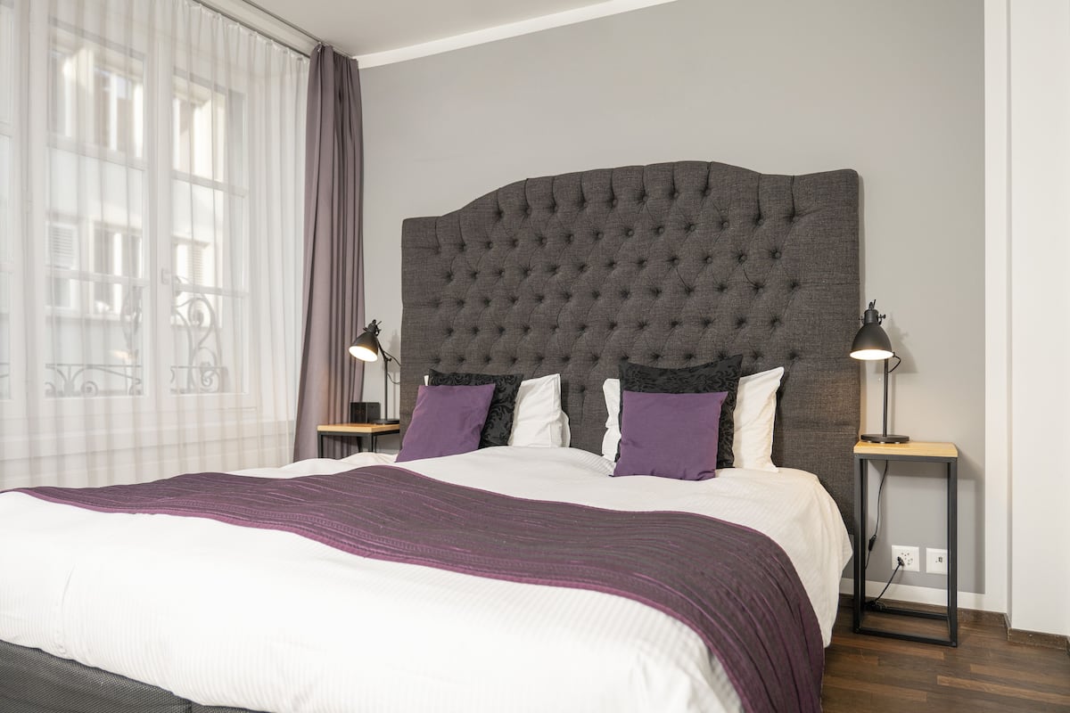 A neatly made bed with a tufted headboard, two bedside tables with lamps, and a window with sheer curtains.