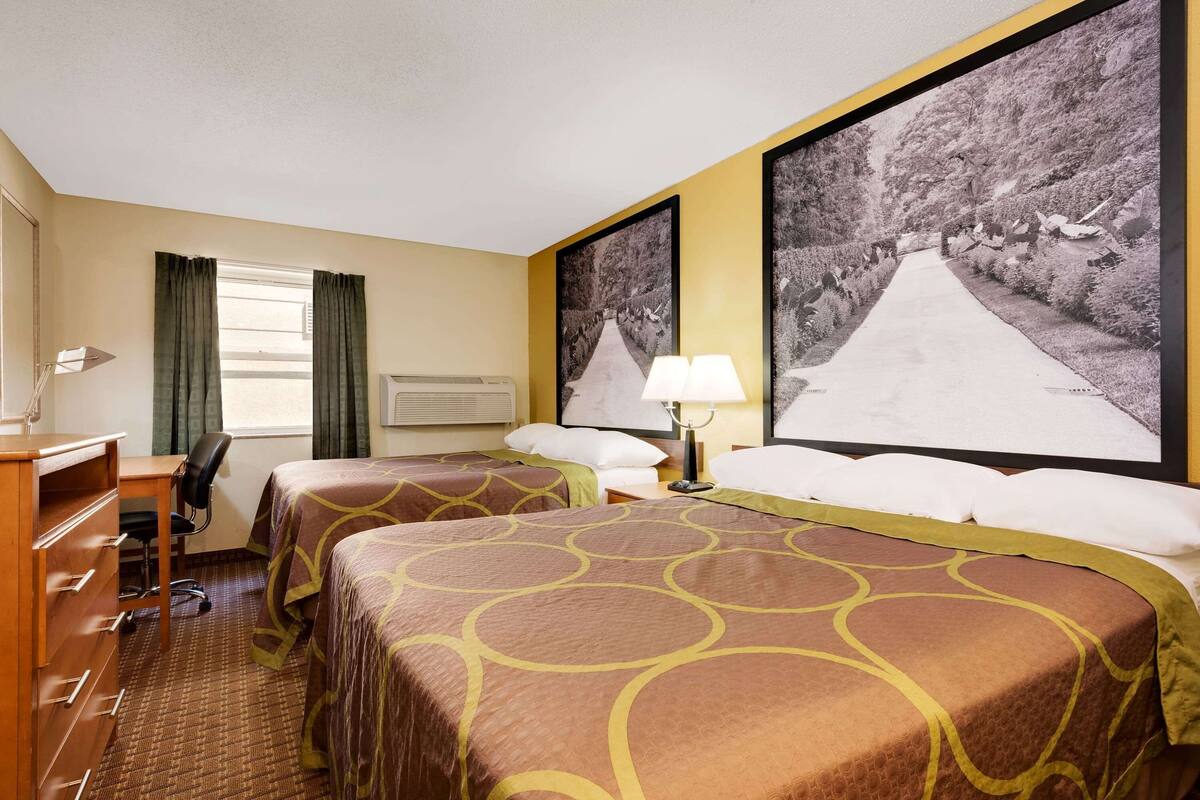 Une chambre d’hôtel avec deux lits, un bureau, une chaise, une lampe et deux grandes images encadrées accrochées au mur.