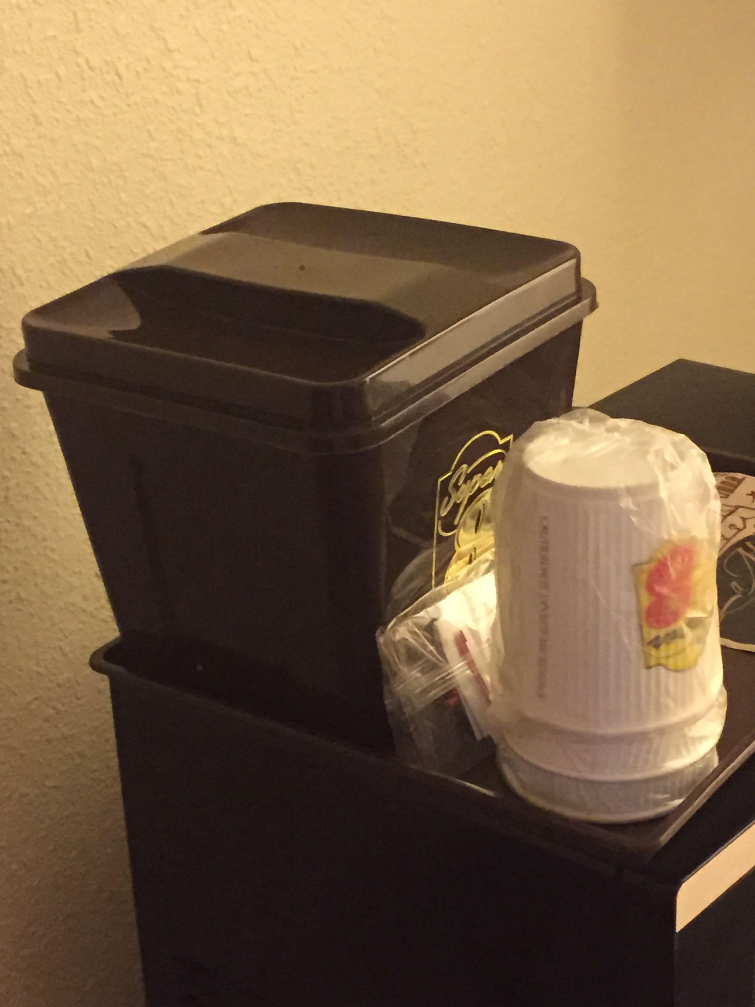 Un bote de basura negro con una bolsa de plástico blanca que contiene una taza blanca con diseño rojo, sobre una mesa negra.