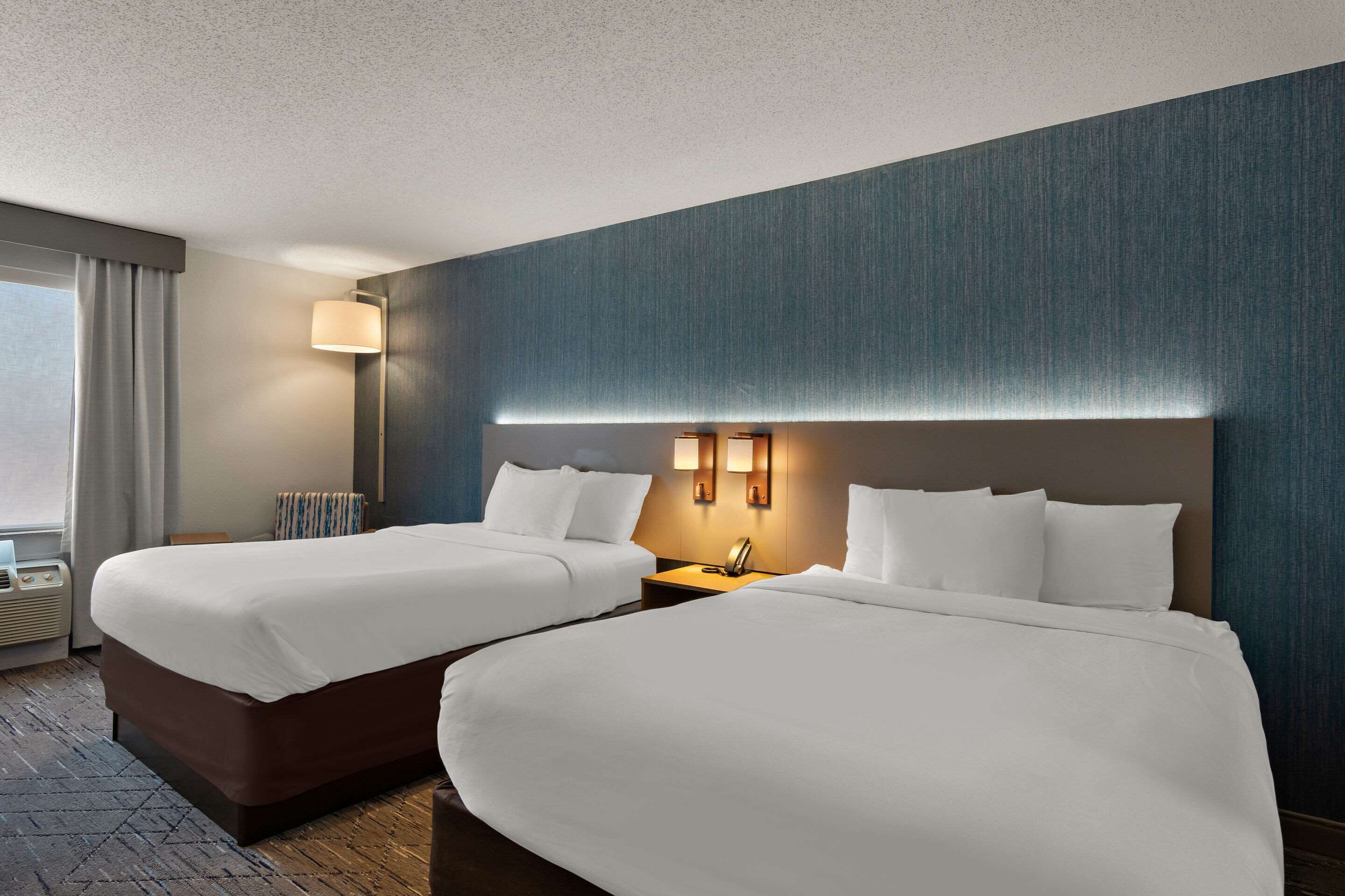 A hotel room with two beds, a nightstand with a lamp, a window with curtains, and a radiator.