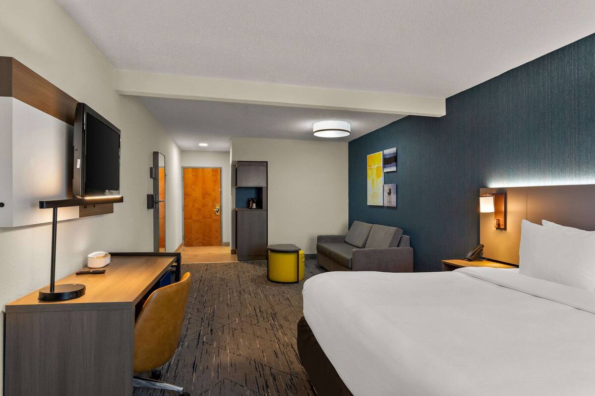 Ett hotellrum med en säng, ett skrivbord, en stol, en tv och ett litet bord med en lampa.