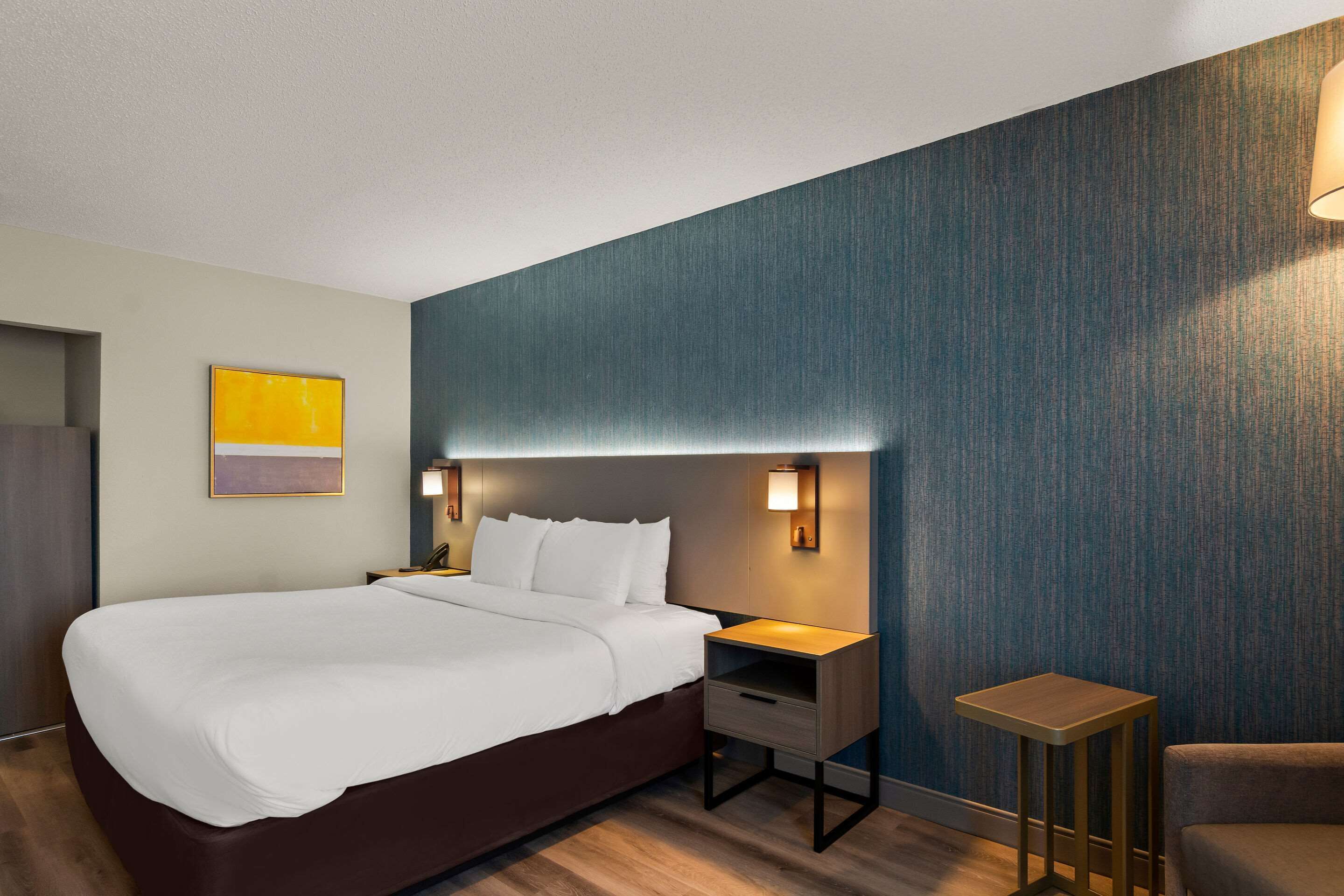 A modern hotel room with a large bed, bedside tables, a small seating area, and a wall-mounted lamp.