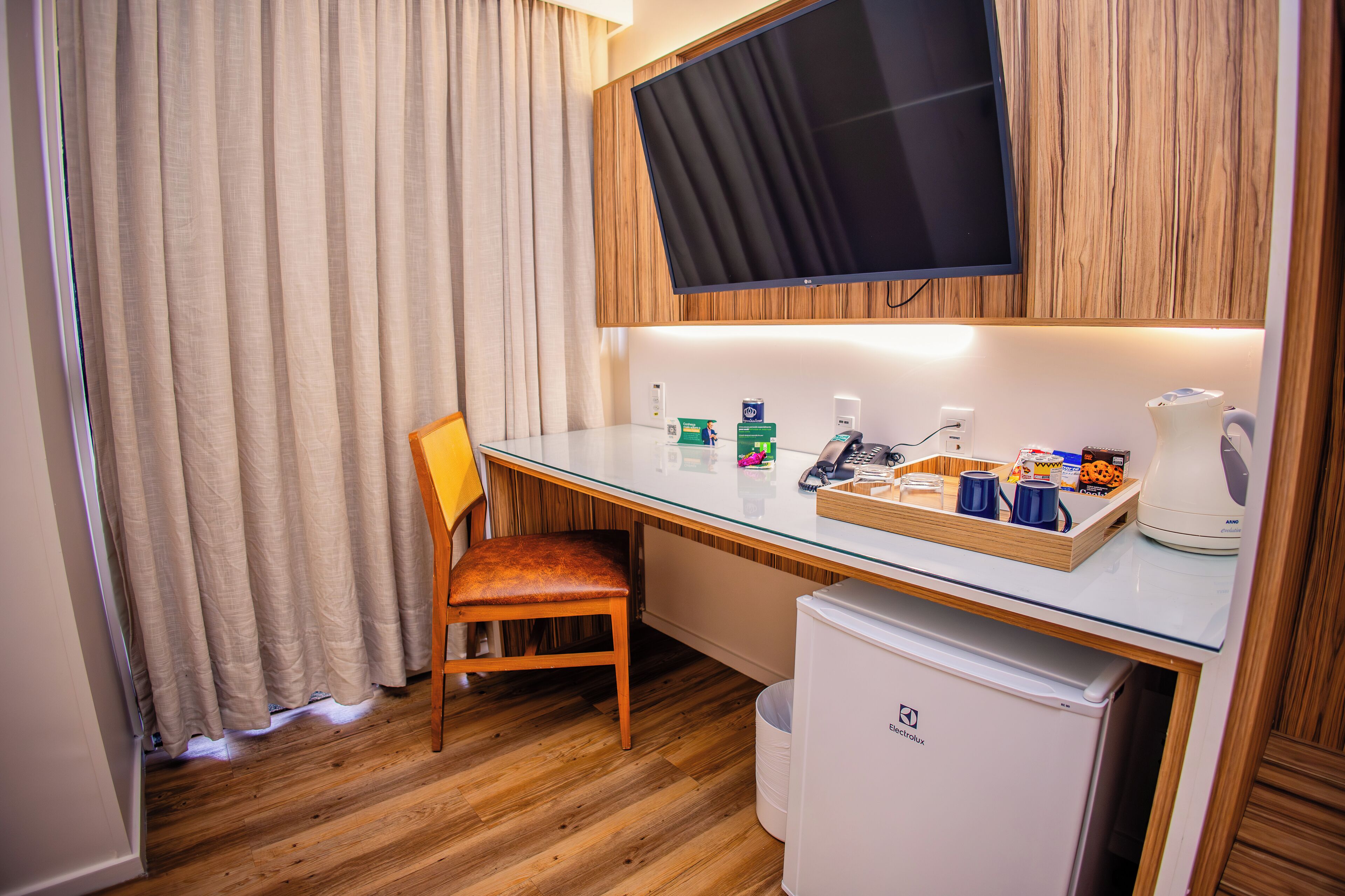 A compact hotel room with a wooden desk, a television mounted above it, a small refrigerator, and a chair.