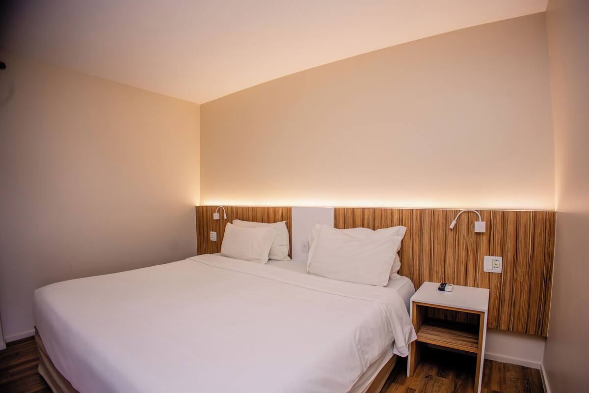 Een opgemaakt bed met witte lakens, een nachtkastje met een lamp en een houten hoofdbord.