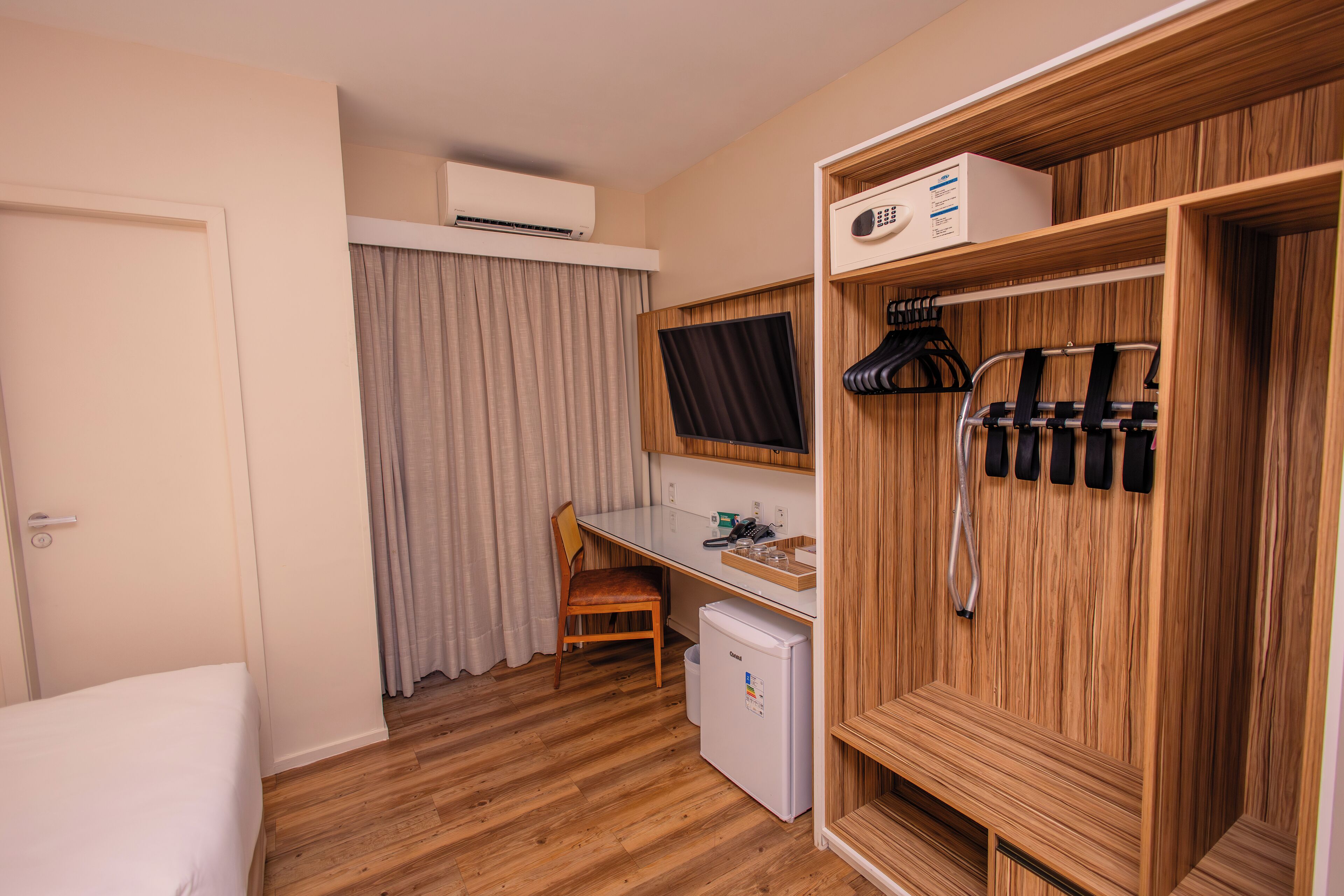 A compact room with a bed, a desk, a chair, a small refrigerator, a wardrobe, and a window with curtains.