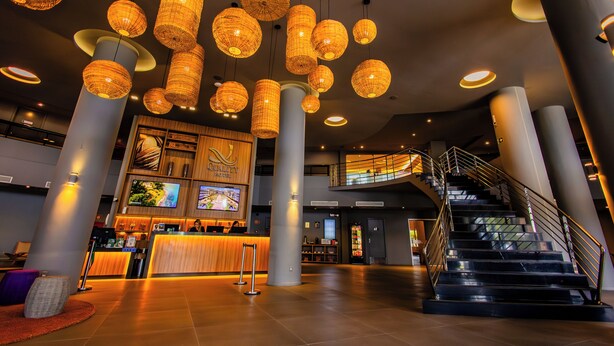 A modern hotel lobby with a reception desk, large columns, a staircase, and hanging wicker lamps.