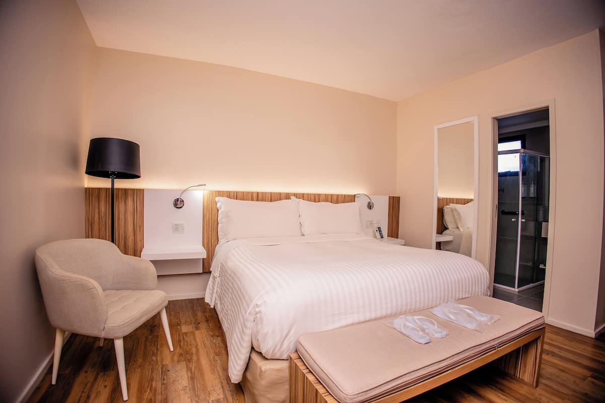 Een moderne hotelkamer met een groot bed, nachtkastje, stoel, bankje, spiegel en een glazen deur naar een douchecel.