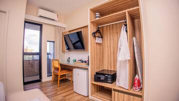 A hotel room with a wooden wardrobe, a flat-screen TV, a mini-fridge, a desk, and a view of the city.