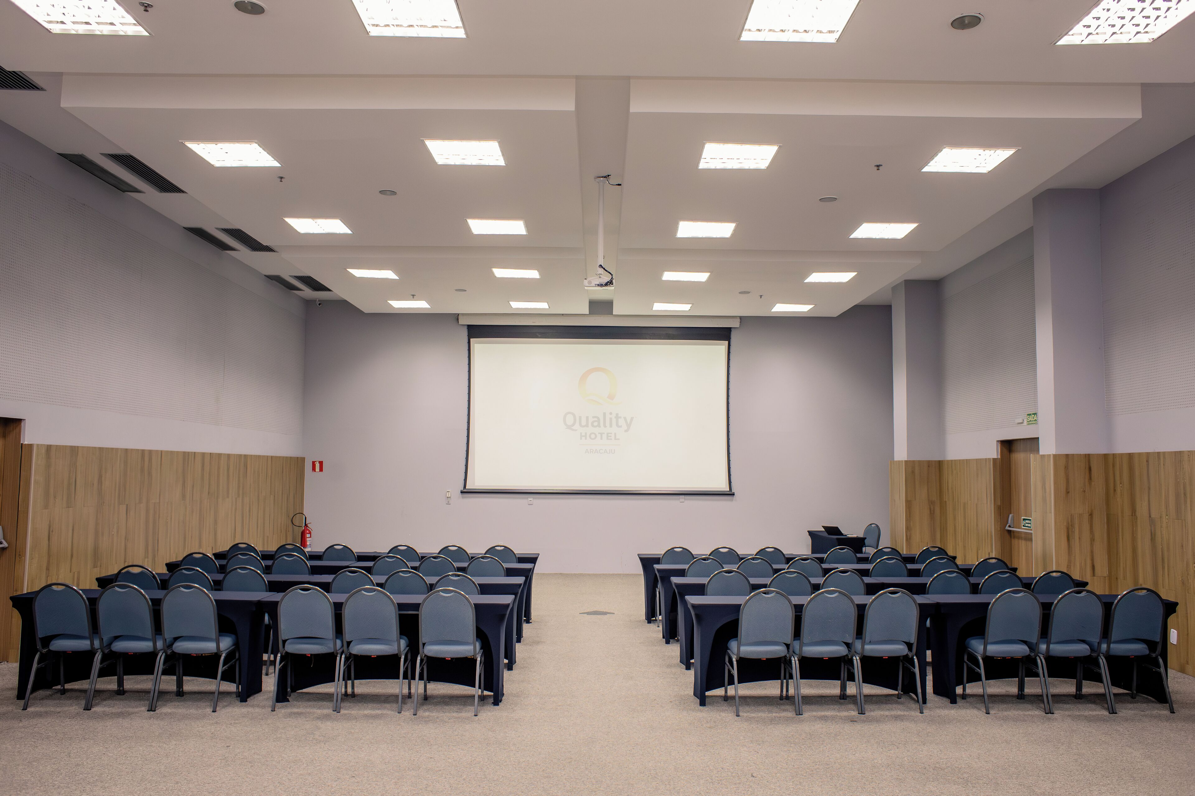 Sala de conferência com tela de projeção exibindo "Quality Hotel" e um extintor de incêndio na parede.