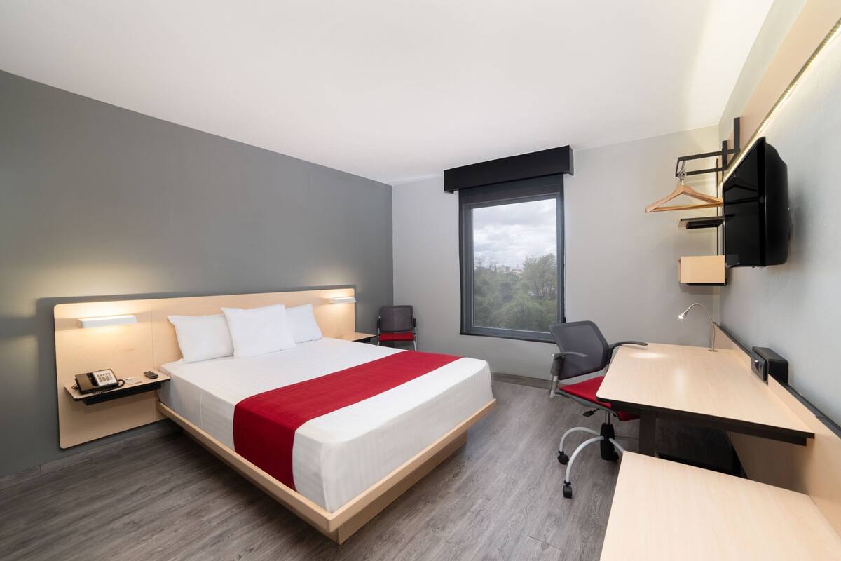 A modern hotel room with a large bed, a desk, a chair, a TV, and a window with a view.