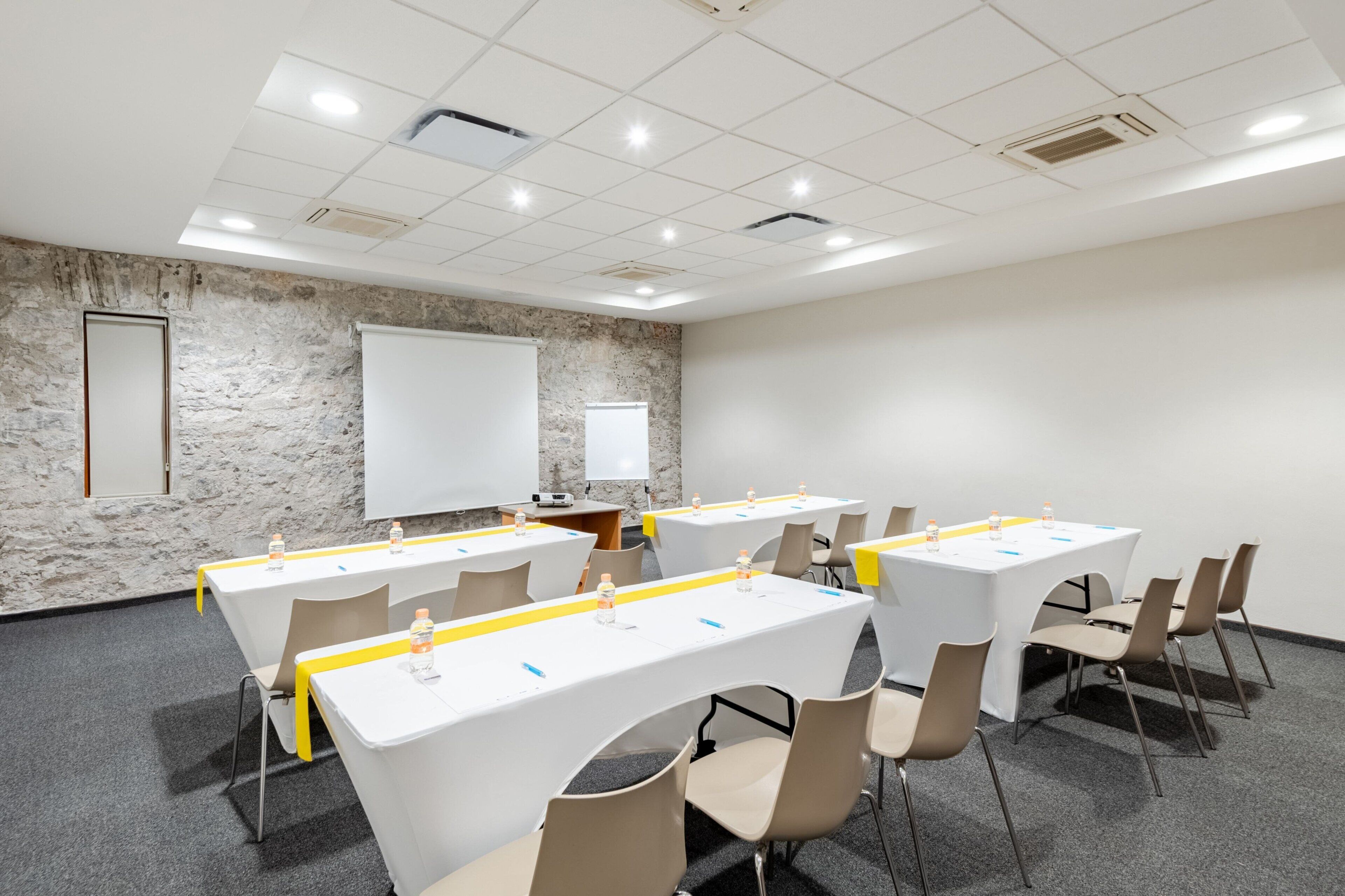 A conference room with white tables, beige chairs, and a projector screen on the wall.