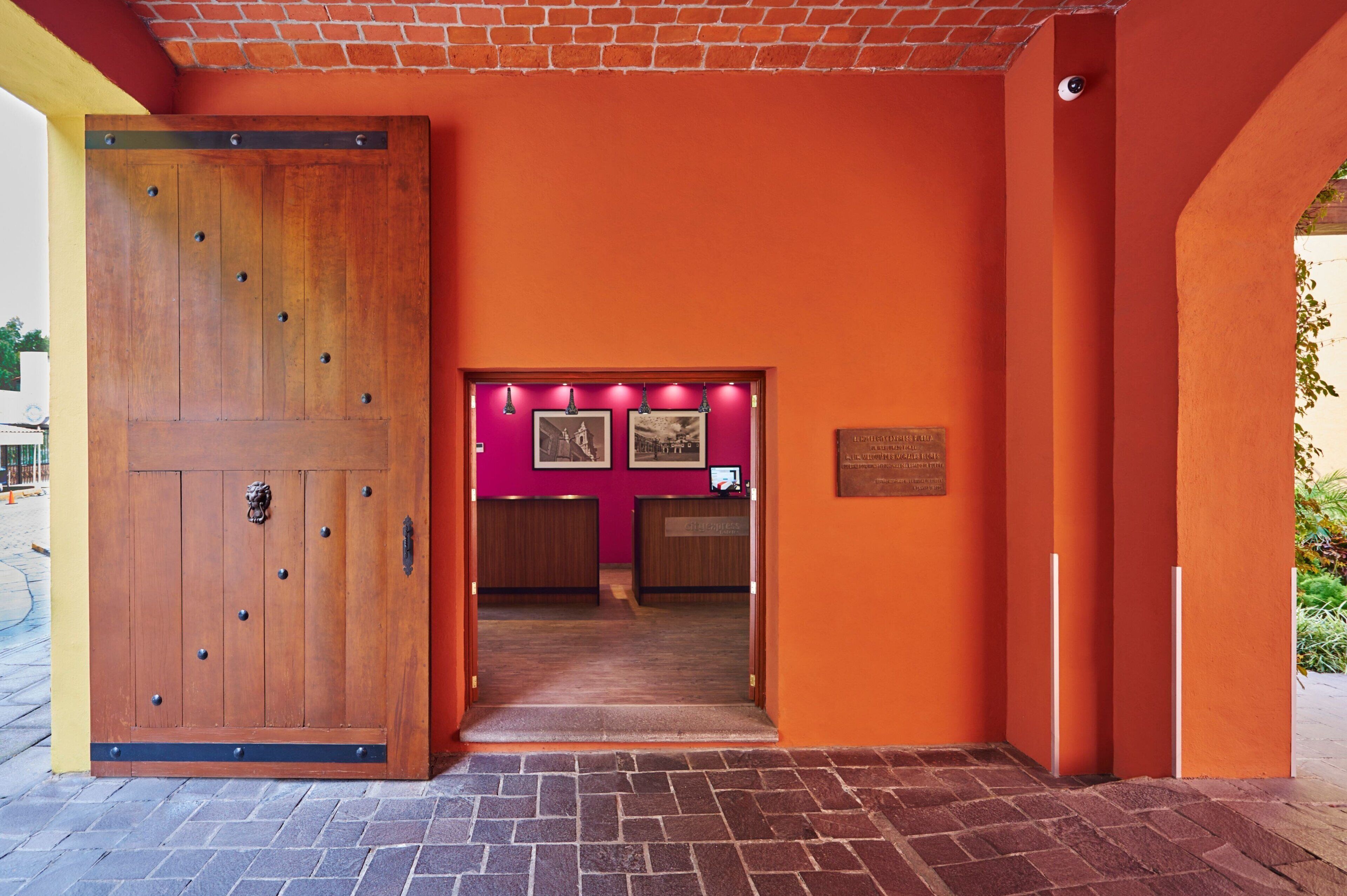 A hotel entrance with a large wooden door, a reception desk, and a pink wall with framed pictures.