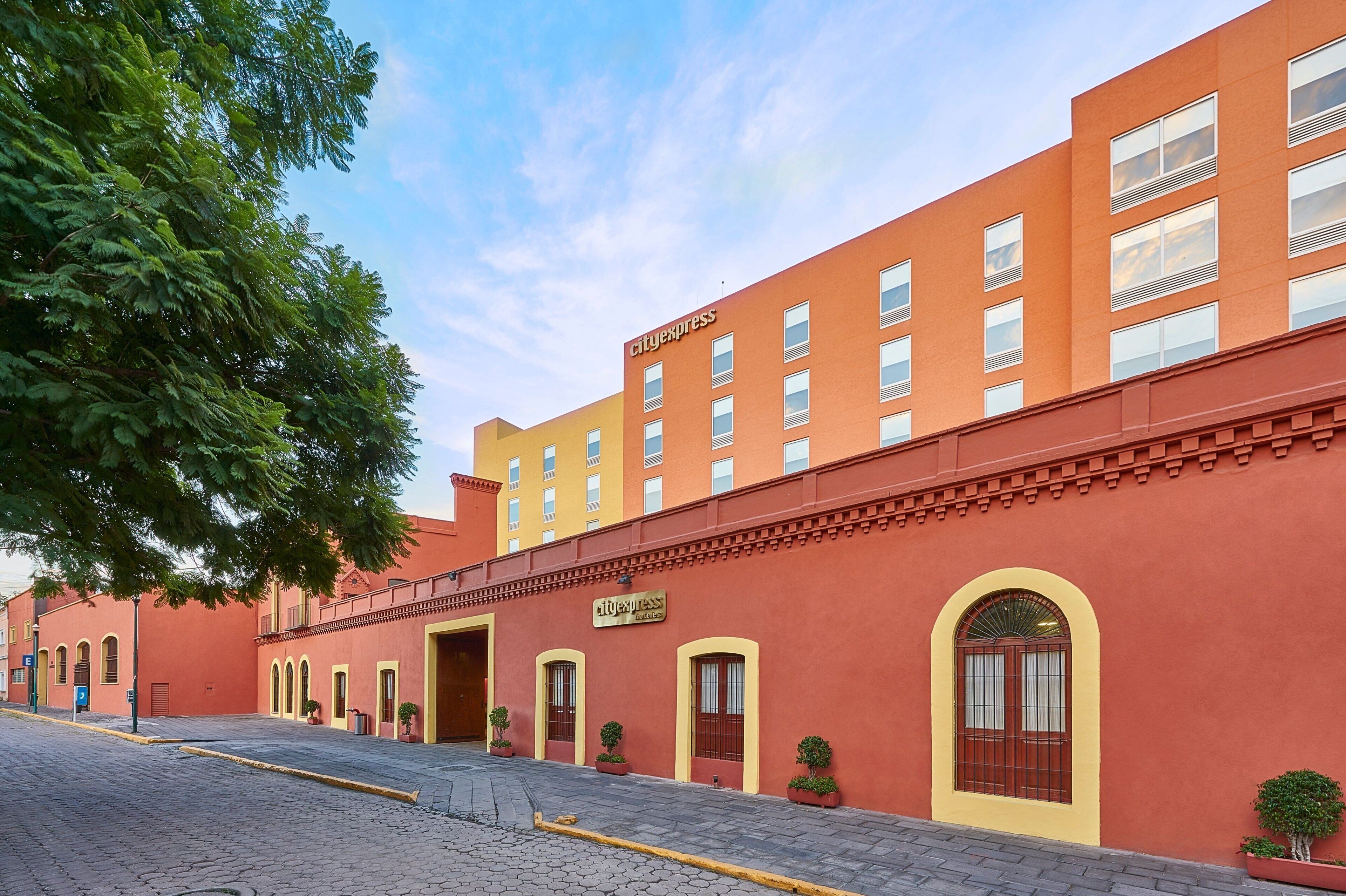 Un edificio rojo con el logotipo de CityExpress, múltiples ventanas y una calle pavimentada con un árbol a la izquierda.