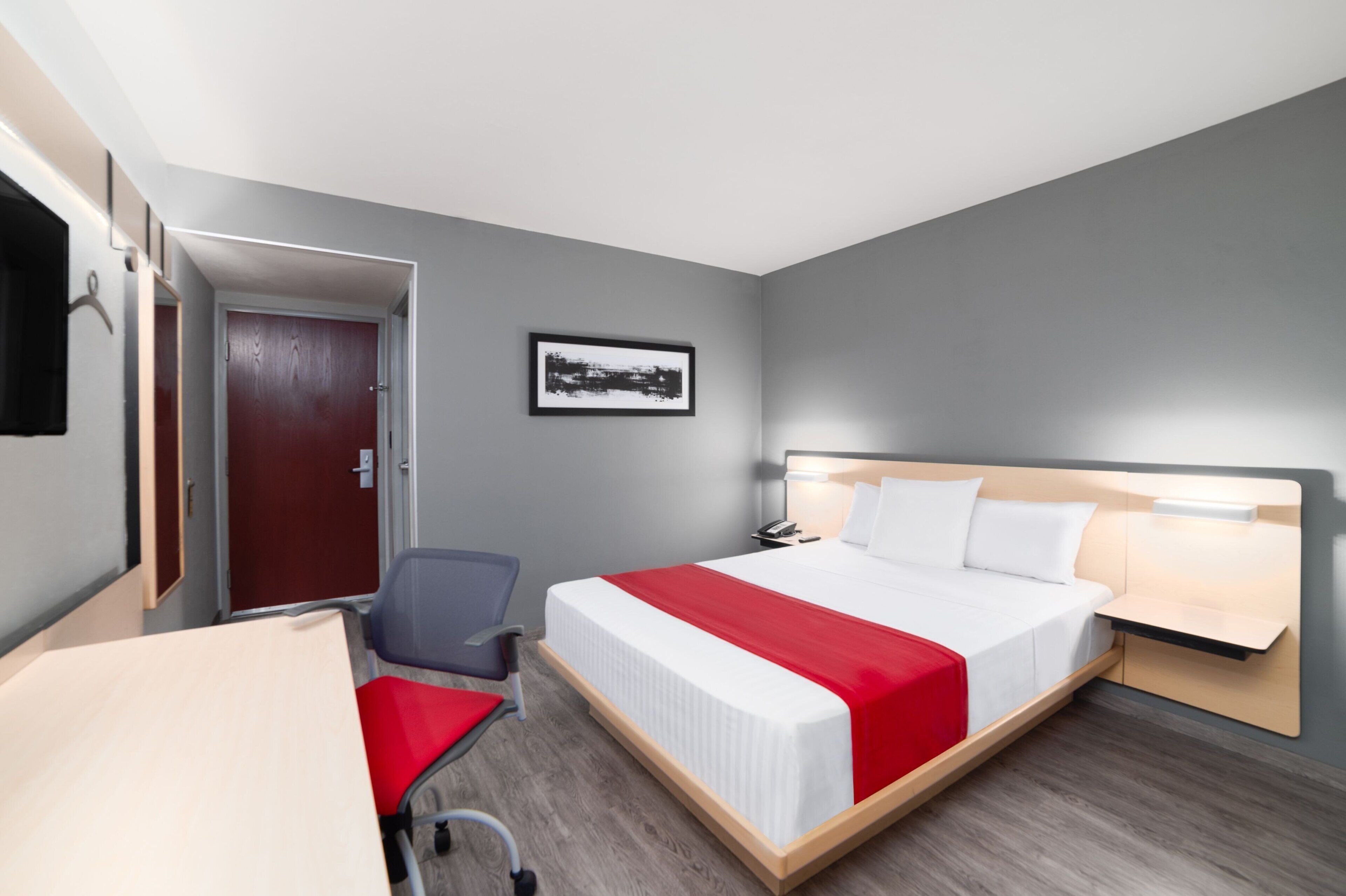 Habitación de hotel con cama, escritorio, silla, puerta y un cuadro en la pared.