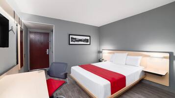 Habitación de hotel con cama, escritorio, silla, puerta y un cuadro en la pared.