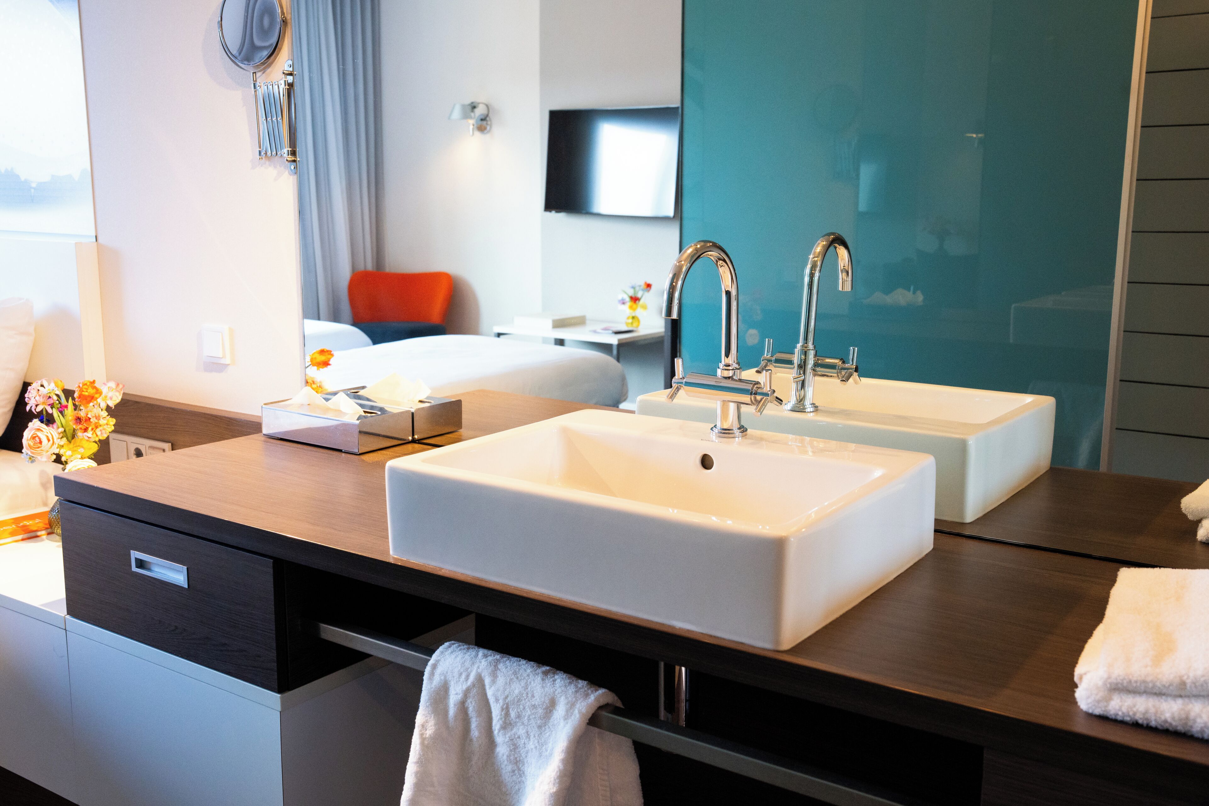 Un baño de hotel con dos lavamanos, un espejo, un perchero para toallas y una cama al fondo.