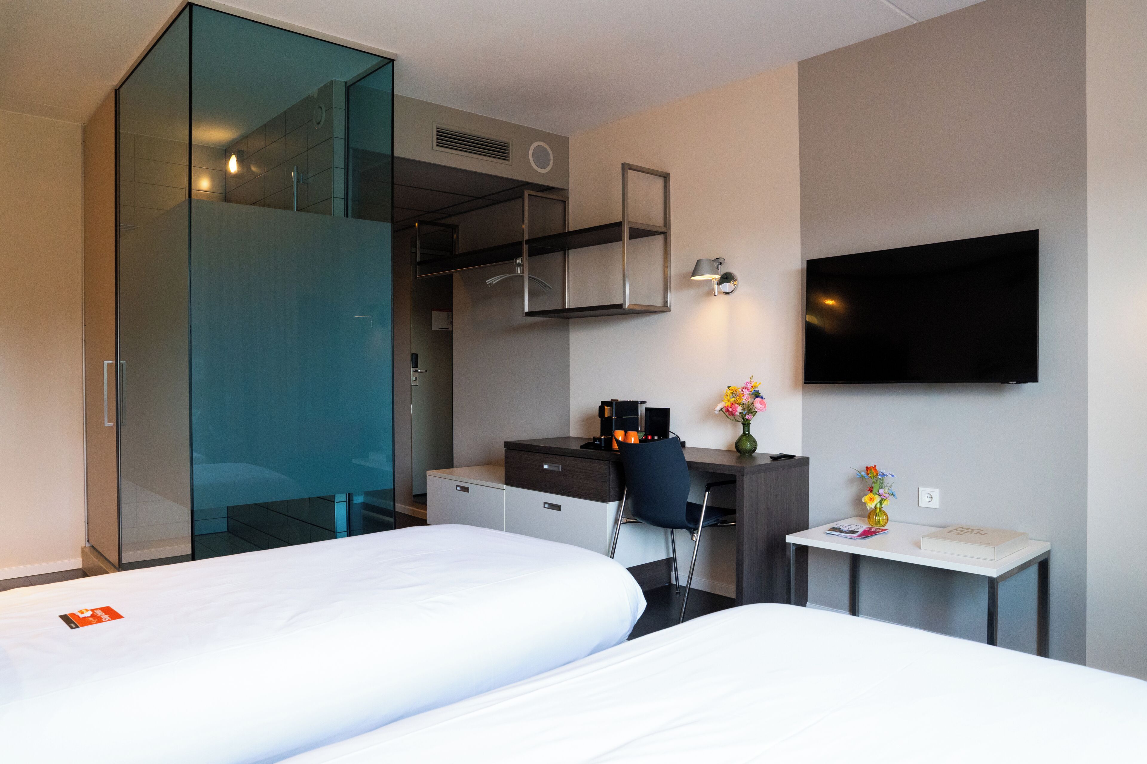 Habitación de hotel con dos camas, un escritorio con silla, televisor y una mesita con un jarrón de flores.