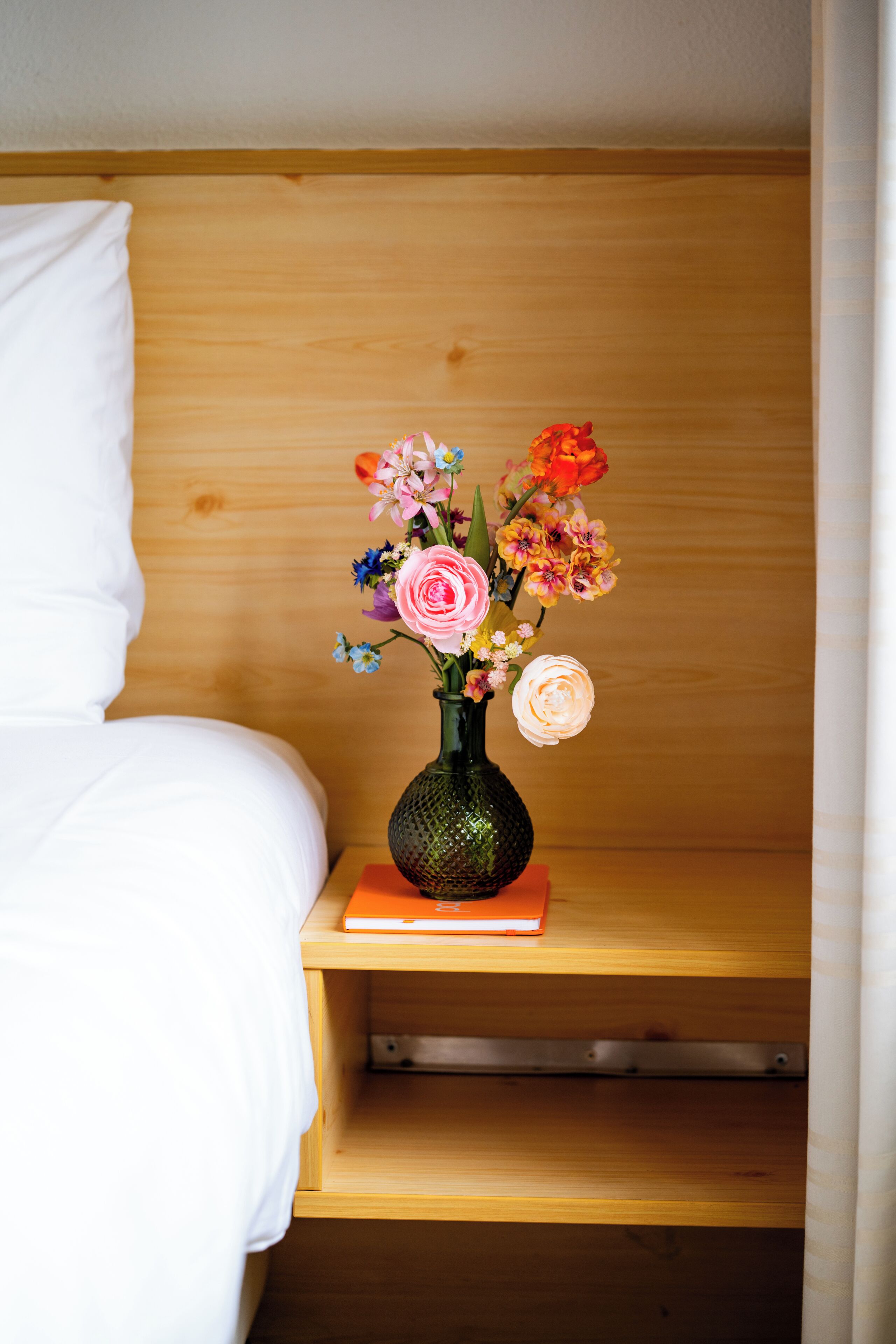 A wooden bedside table with a vase of flowers and a book.