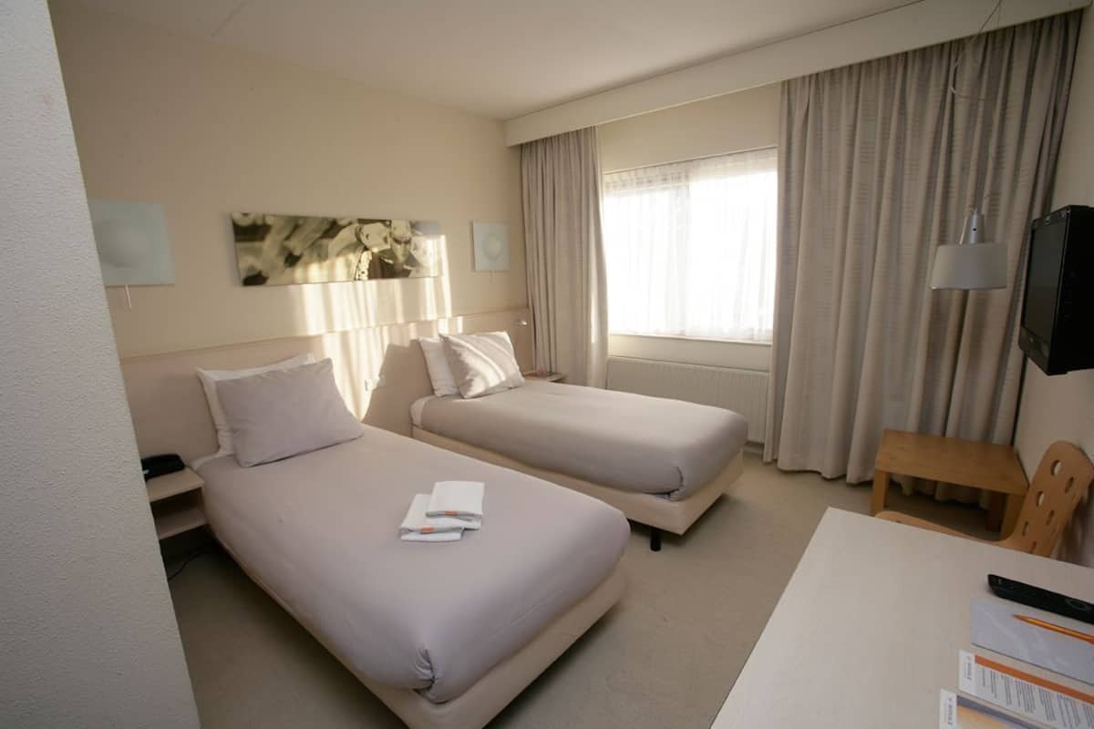 A hotel room with two beds, a desk, a chair, a TV, and a window with curtains.