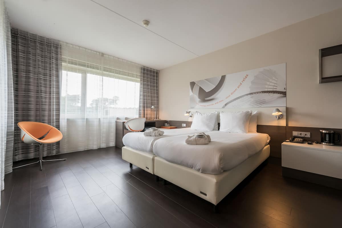 A modern hotel room with a large bed, a chair, a desk, and a view of the city through the window.