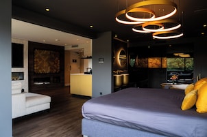 A modern bedroom with a large bed, a seating area, and a kitchen in the background.