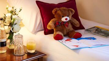 A bed with a red pillow, a teddy bear, a book, a glass of orange juice, and a vase with white flowers.