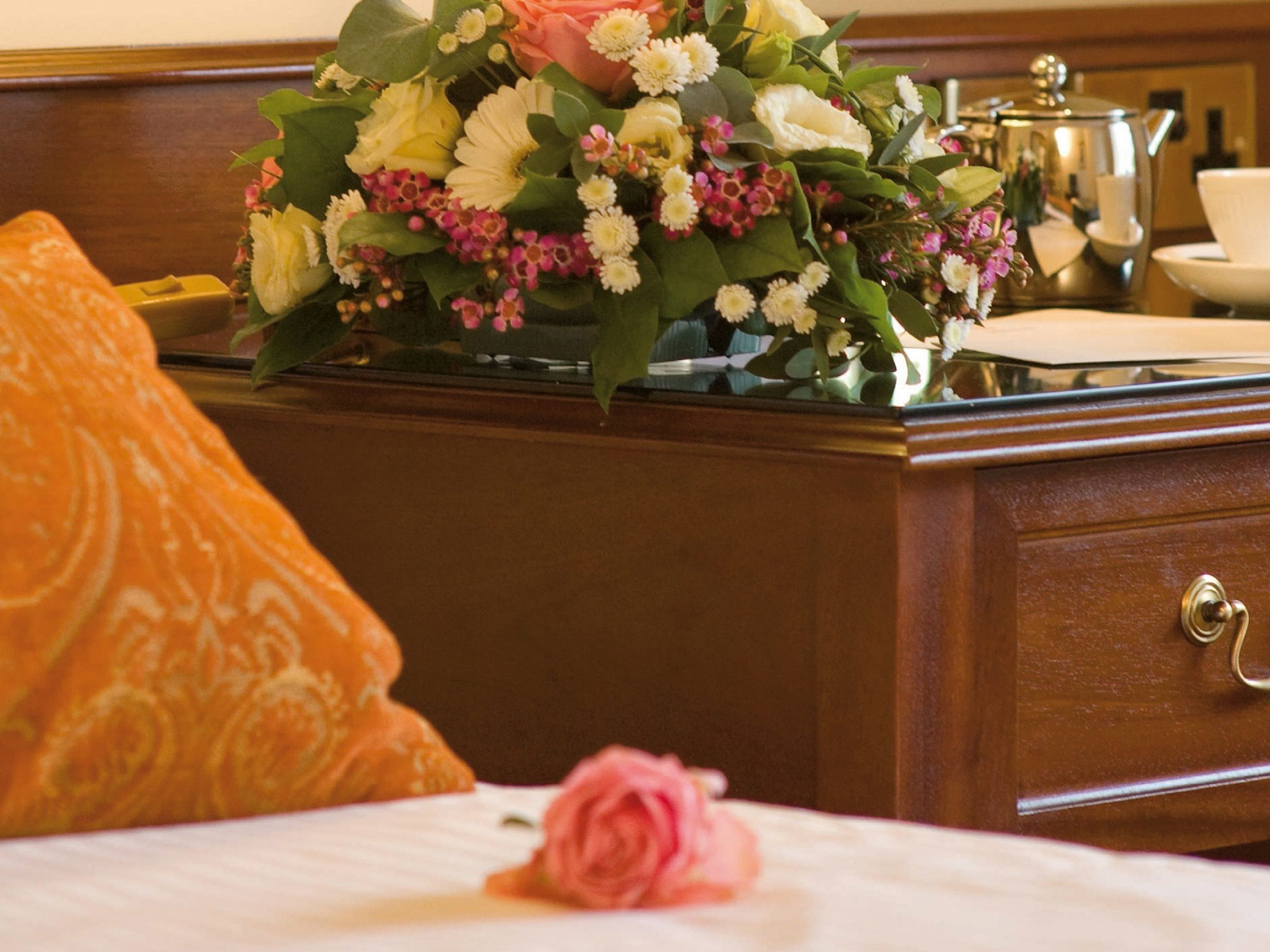 Ein Hotelzimmer mit einem Holzkapmierer, einem Bett mit einem orangefarbenen Kissen mit Muster und einer Blumenkomposition auf dem Kapmierer.